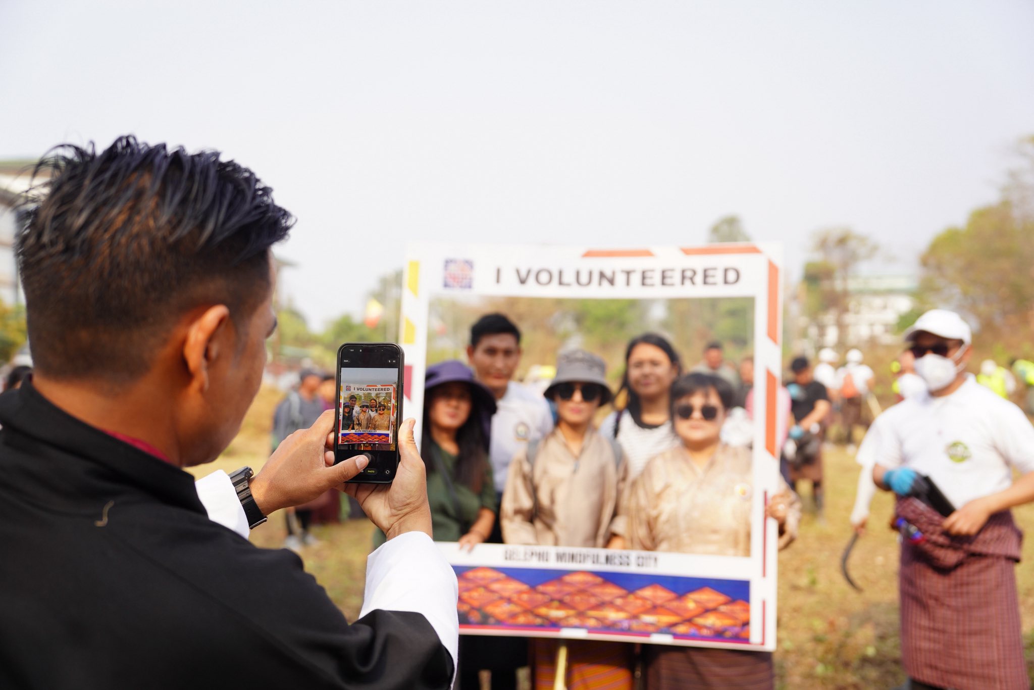 Volunteers pose in an "I Volunteered" photo frame at a Gelephu Mindfulness City voluntary zhabtog programme, one filming on a phone, another holding a brush-clearing sickle